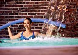 Eine Frau lächelt mit einer Schwimmnudel in den Händen, umgeben von sprudelndem Wasser in einer Therme von Bad Füssing.