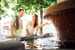 Ein glückliches Paar in Freizeitkleidung genießt die Urlaubsatmosphäre am Wasserbrunnen in einem Park in Bad Füssing.